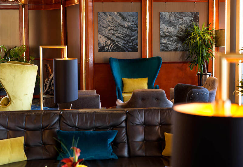 Cozy hotel lounge with leather sofa, teal velvet wingback chair, colorful pillows, wood paneling, modern lamps and plants.
