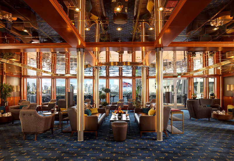 Warm hotel lounge with plush seating, mirrored ceiling, wood accents and nautical carpet, large windows with river views.