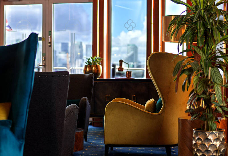 Cozy hotel lounge with velvet armchairs, plants and warm lamp by large windows showing a blurred city skyline