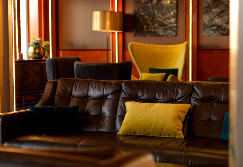 Cozy hotel lounge with tufted leather sofa, yellow accent pillows, velvet wing chair, warm wood panels and ambient lighting.