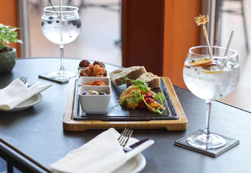 Tapas sharing board with olives, dips, roasted peppers and bread on a wooden tray, served with two gin-and-tonic cocktails.