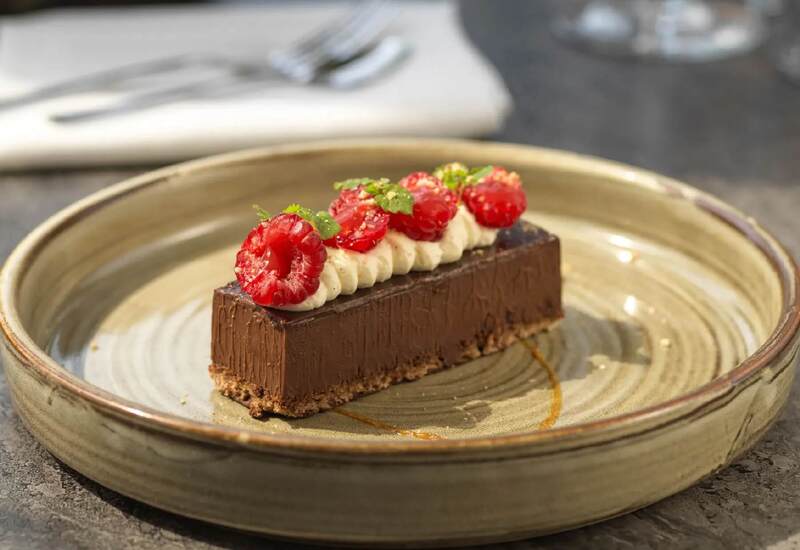 Decadent chocolate tart with piped cream, fresh raspberries and mint on a rustic ceramic plate in hotel dining.