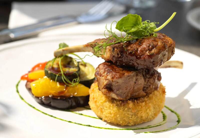 Two glazed lamb chops stacked on a golden breadcrumb croquette, served with grilled vegetables and pea-tendril garnish.