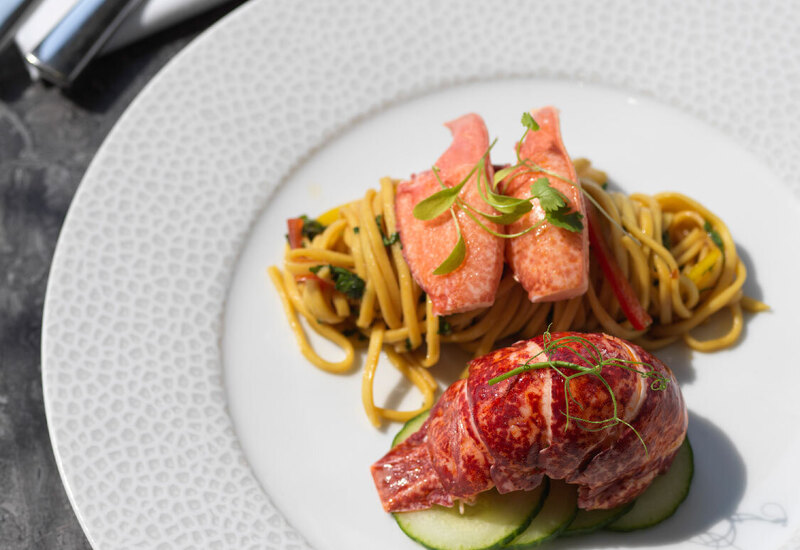 Plated lobster tail with tossed noodles, cucumber slices and microgreens on a textured white dinner plate, fork and knife.