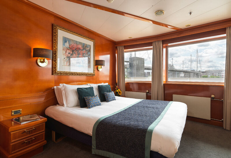 Wood-paneled double room with king bed, patterned cushions, framed art and bedside lamps; windows with modern bridge and city view.