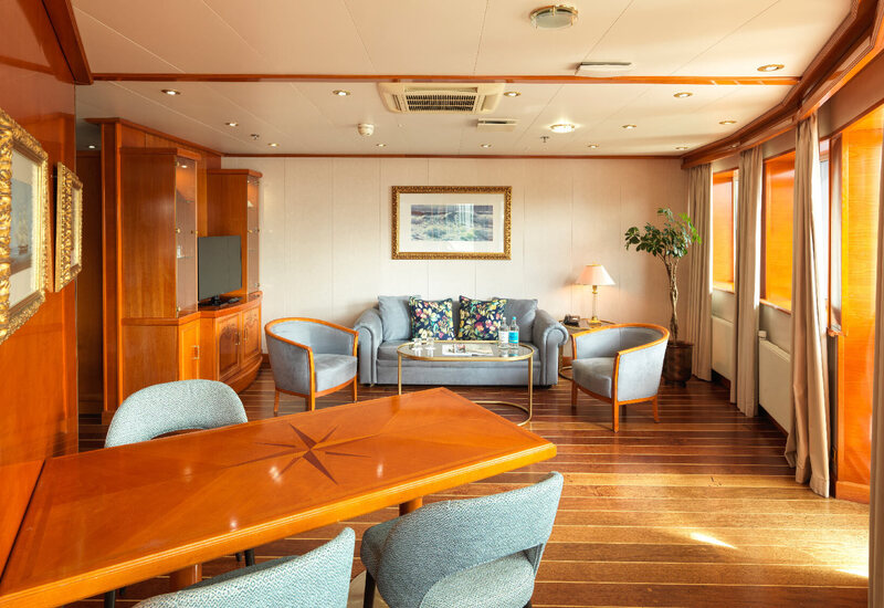 Luxury suite lounge with warm wood finishes, dining table, plush sofa and chairs, coffee table, lamp and sunlit windows.