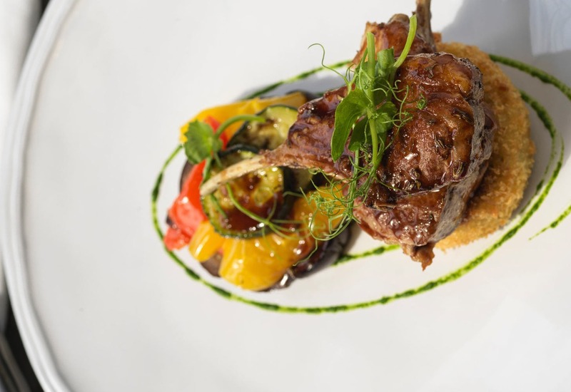 Seared lamb chops with herb glaze on crispy crumb cake, grilled peppers and zucchini, microgreens and green herb oil swirl on white plate
