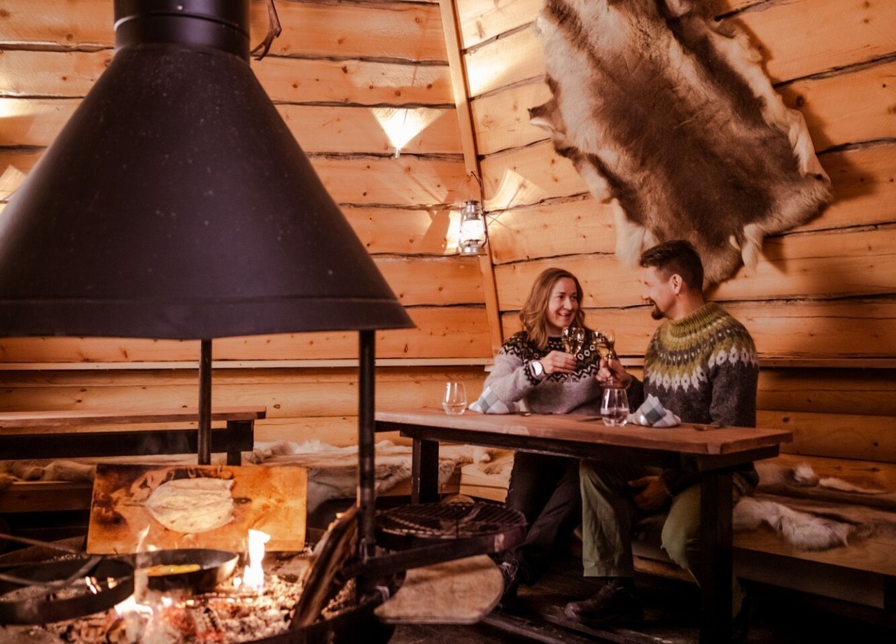 A couple sitting at a table in a wooden hut with a fireplace in the middle.
