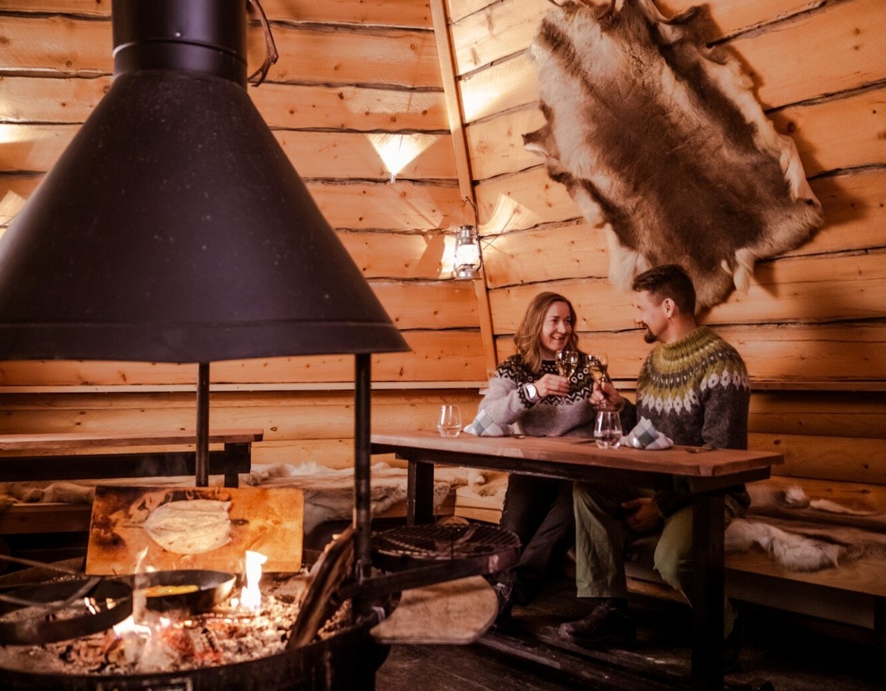 A couple sitting at a table in a wooden hut with a fireplace in the middle.