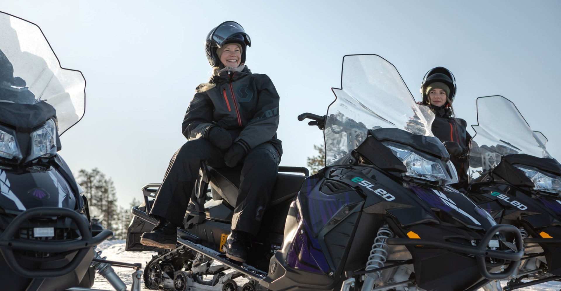 A woman sitting sideways on an electric snowmobile and laughing.