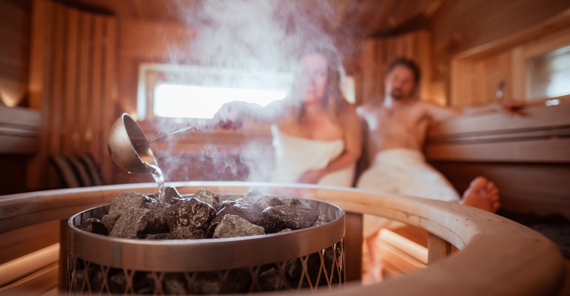 A couple in the steam sauna, with the woman pouring water on the coals.