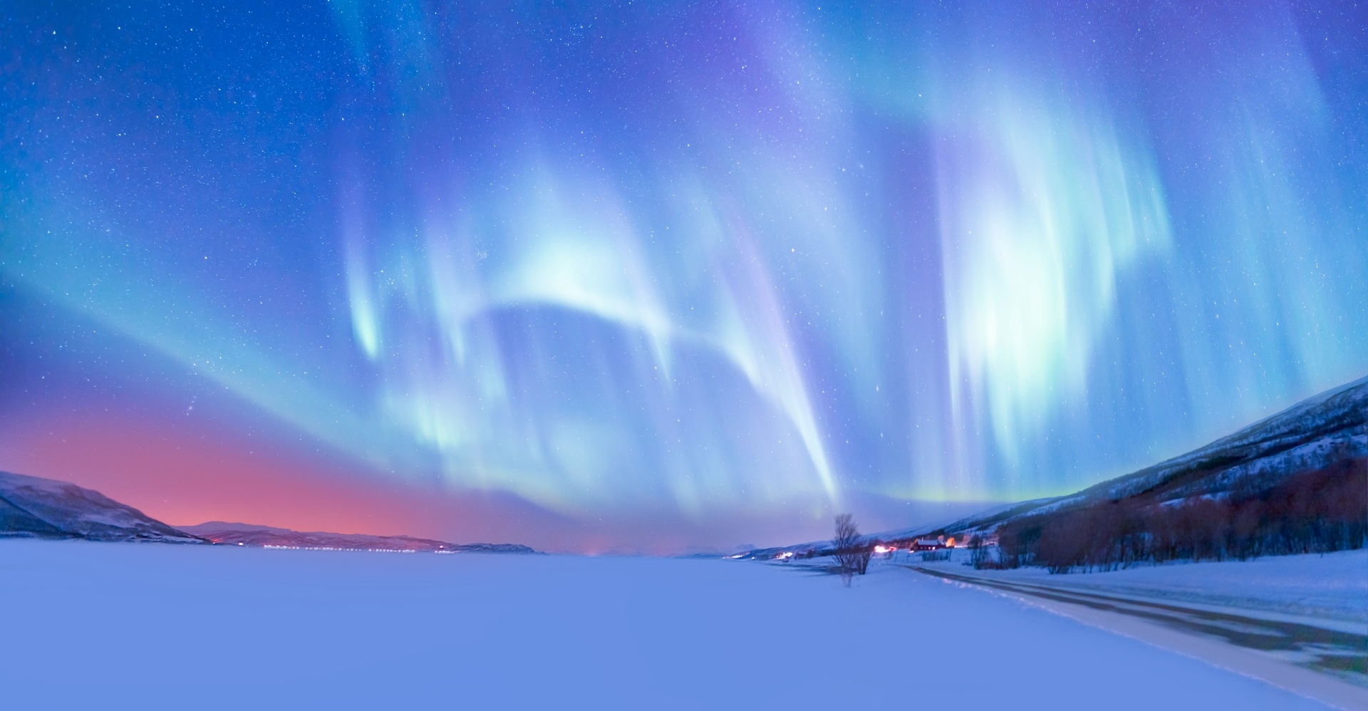 Northern lights above a frozen lake, with a small village on its right bank.