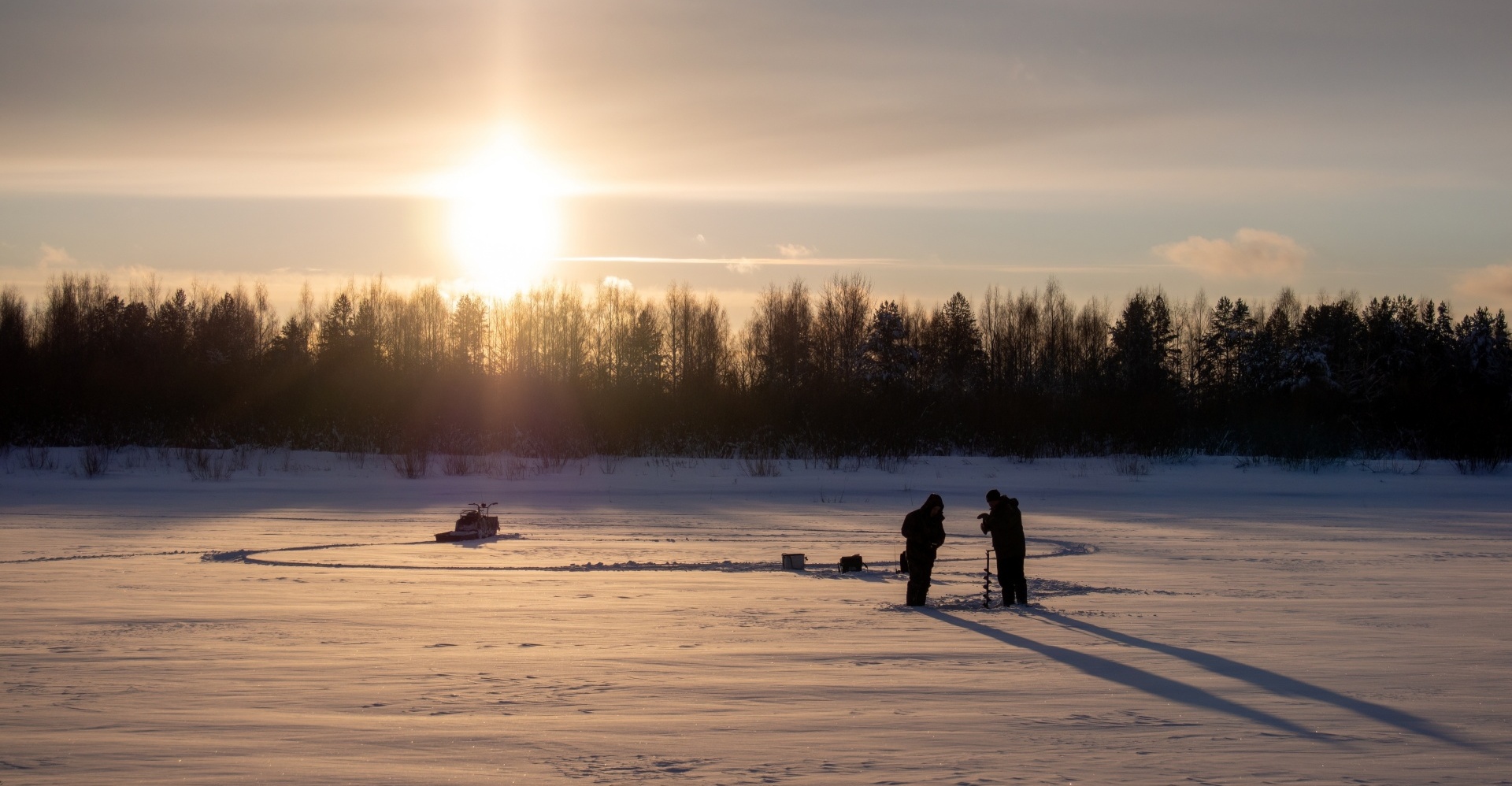 Two people ice fishing under the sunset.