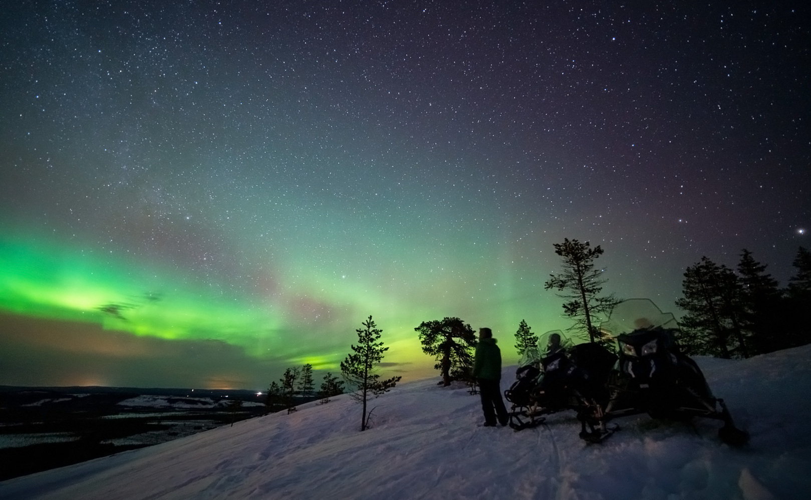 Green and blueish northern lights visible above a snowy hill.