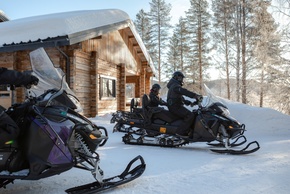 木造ロッジ前の雪上バイクに乗る宿泊客、雪に覆われた松林での冬のアクティビティ