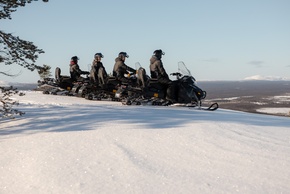 Tour en motos de nieve: cuatro huéspedes con casco en fila sobre una cima nevada, vistas panorámicas de bosque y montañas
