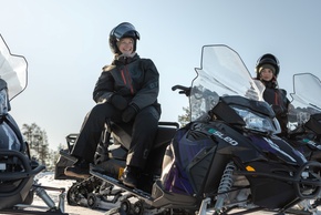 ヘルメットを着けた2名のゲストが雪上バイクに乗り、冬装備で晴れた雪景色と松林を背景にしている。