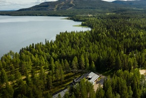 Alojamiento tipo lodge con techo metálico junto a un lago, rodeado de bosque de pinos y senderos, vista a montañas