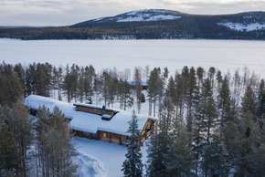 A snow-covered timber lodge surrounded by trees, with other lodges and the lake in the background.