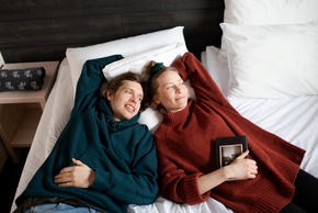 Casal relaxando em cama grande com roupa de cama branca num quarto acolhedor estilo chalé; mulher segura um livro, manta dobrada no criado-mudo