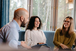 Trois invités souriants discutent dans un salon moderne avec grandes fenêtres et lumière naturelle.