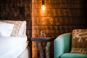 Cozy hotel room corner with rustic wood wall, soft bed, teal armchair, warm pendant light, and natural wood side table