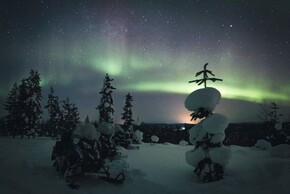 Snowmobiles lined on a snowy clearing with snow-topped pines under a green aurora and starry night sky