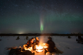 Group around a campfire on a snowy plain beneath a starry sky and green northern lights, with a snowmobile at the edge