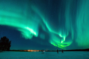 Green aurora borealis over a snowy frozen lake, silhouetted photographers and warm village lights on the horizon