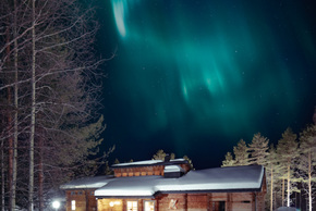 Snow-covered wooden lodge with warm-lit windows among pine trees, beneath vibrant green northern lights