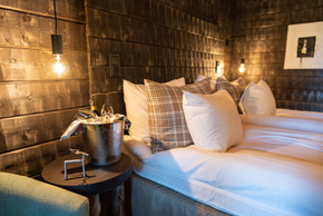 Rustic wood-paneled hotel bedroom with warm hanging bulbs, plush white linens, plaid pillows and champagne on ice at bedside.
