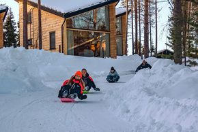 Cabañas de madera modernas con grandes ventanales junto a un camino nevado donde huéspedes se deslizan en trineo entre pinos