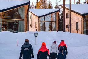 Família puxando trenós coloridos em direção a chalés de madeira modernos numa floresta nevada ao pôr do sol.