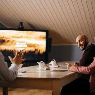 Two people at a wooden table talking lively. In the back, a TV with a presentation.