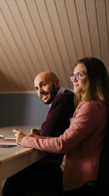 Two people at a wooden table talking lively. In the back, a TV with a presentation.