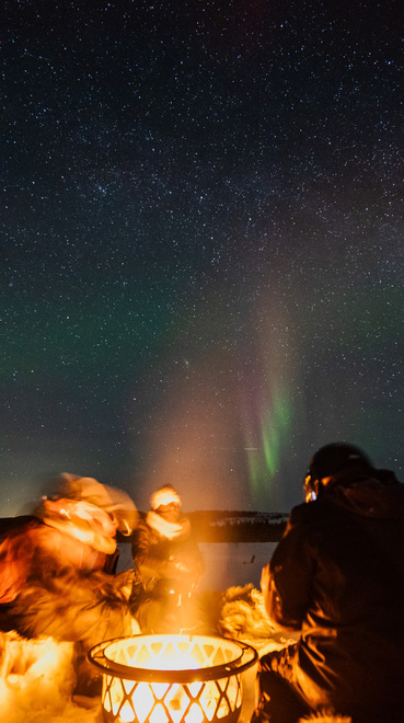 Guests bundled in fur wraps around a glowing outdoor fire pit under a starry sky and green aurora over snowy landscape.