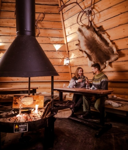 A couple sitting at a table in a wooden hut with a fireplace in the middle.