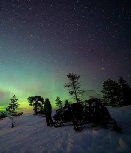 雪の尾根に停めたスノーモービルと、星空に広がる緑のオーロラ