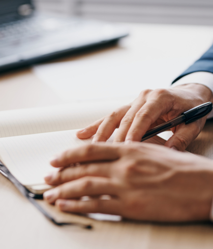 Man's hands writing in a notepad, with a laptop sitting on his right side.