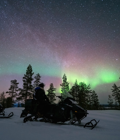 雪地摩托上的游客在松林前观赏绿色北极光与满天繁星，夜晚雪地景致。