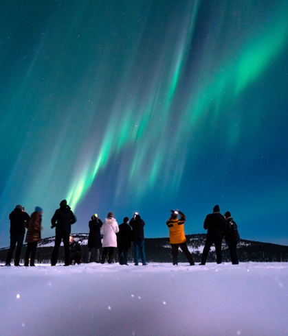 Huéspedes en tour de auroras: grupo en la nieve fotografiando cortinas verdes del norte sobre un horizonte montañoso.