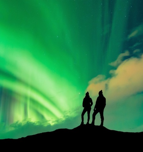 Two silhouettes on top of a hill admiring the northern lights in front of them.