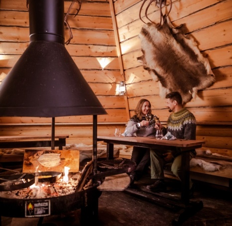 A couple sitting at a table in a wooden hut with a fireplace in the middle.