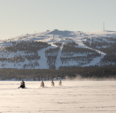 People riding snow scooters, with a large hill coated in snow and covered in trees behind them.