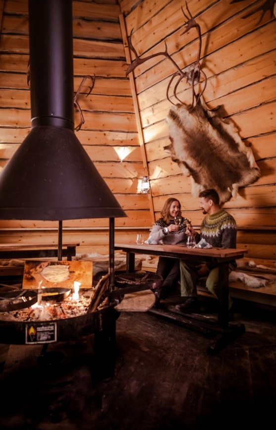 A couple sitting at a table in a wooden hut with a fireplace in the middle.