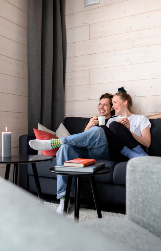 A couple cuddling on the couch and laughing. They hold teacups and look out the window.