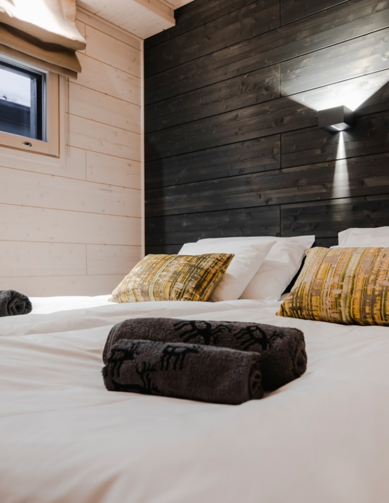 A queen size bed, with black wooden wall behind and an elegant lamp mounted to it.