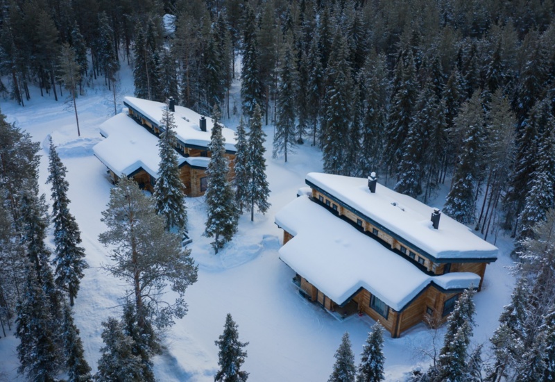 Two wooden lodges covered in snow and surrounded by trees.
