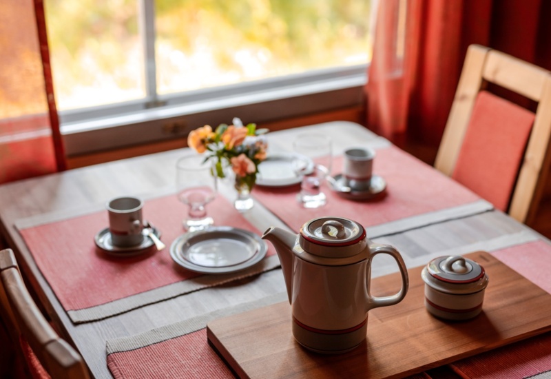 A teapot placed on a small square table decorated with flowers and red placemats.