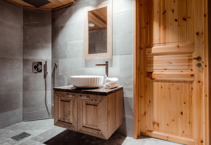 A bathroom with stone tiles, with a sink placed in the middle on top of a wooden cupboard.